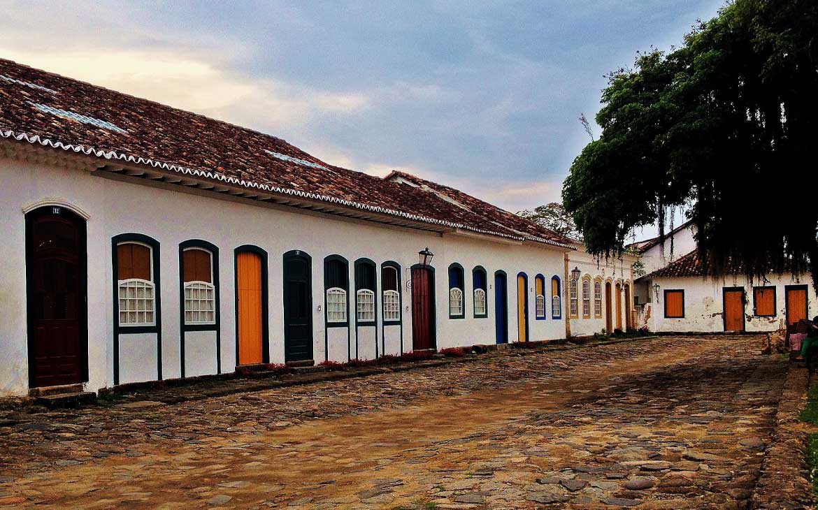 Foto, durante o dia, de casas brancas com janelas e portas coloridas, em rua de pedras de Paraty.