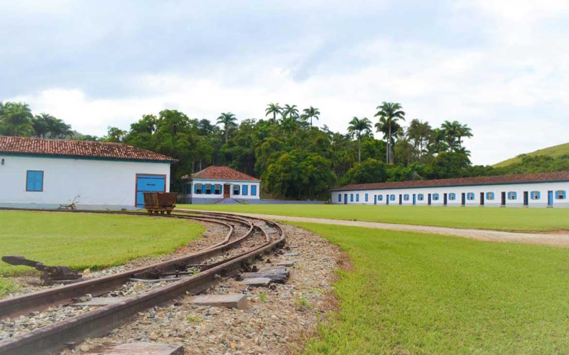 Foto, durante o dia, de trilho de trem em frente a casas brancas com janelas azuis e telhas de madeira.