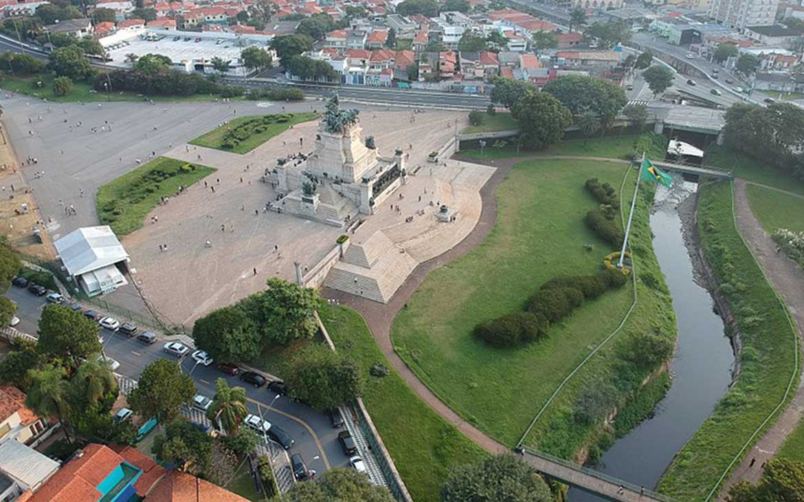 Imagem aérea, durante o dia, de carros passando em avenida ao lado de praça onde há monumento à Independência do Brasil, formado por esculturas em granito e bronze, localizada às margens do Riacho do Ipiranga
